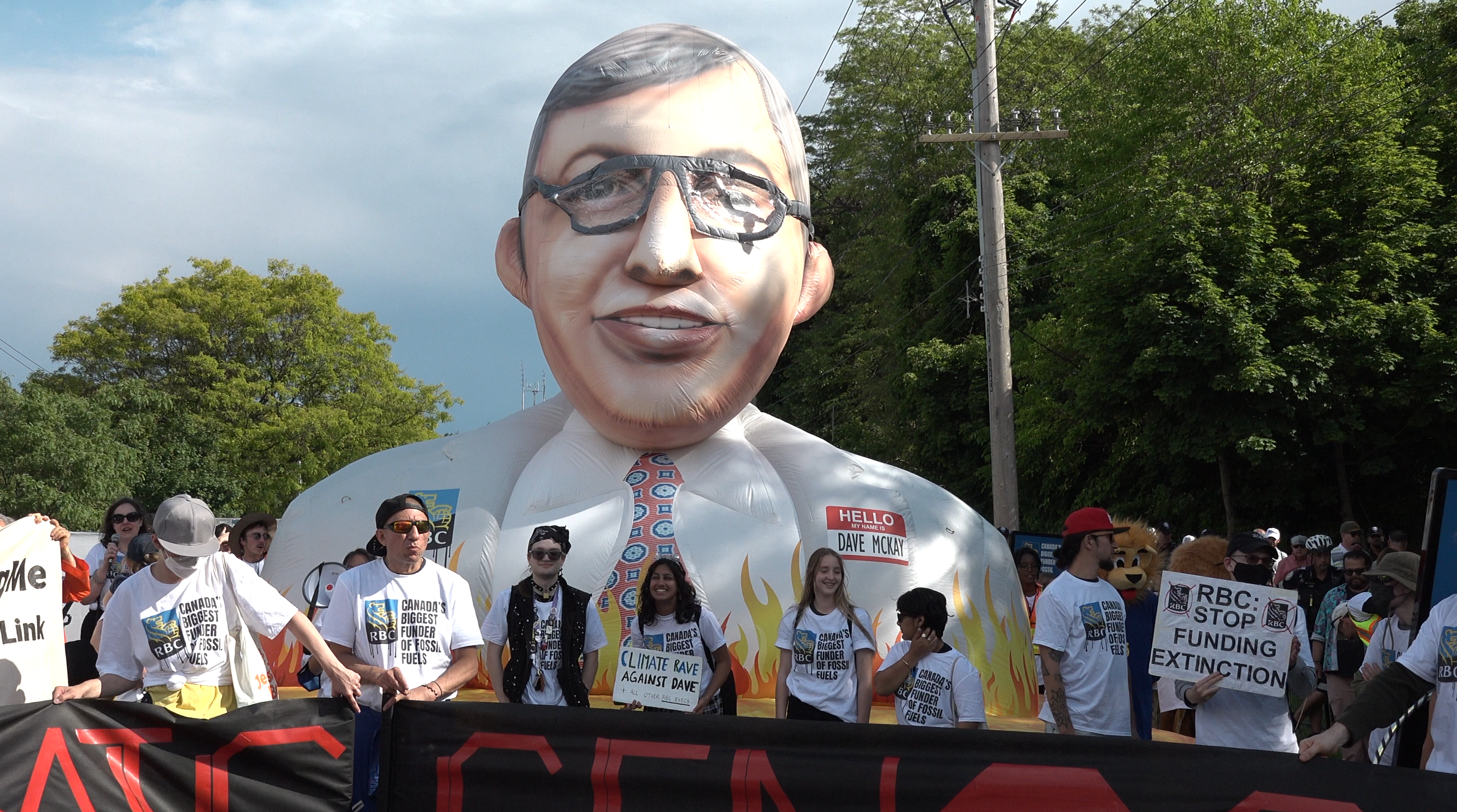 Protestors hold signs and stand in front of a tall inflatable of Dave McKay.
