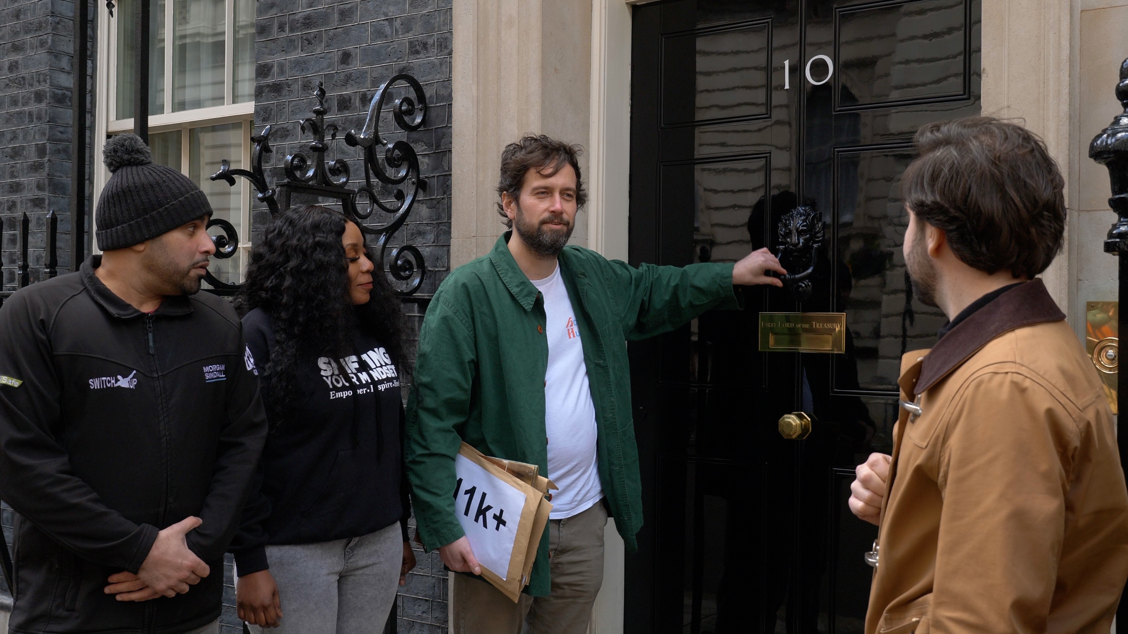 A group of four diverse people stand outside a black door with the number 10 on it. A man in a green shirt holding an envelope with 11k+ written on it lifts the door knocker as the others looks towards him.
