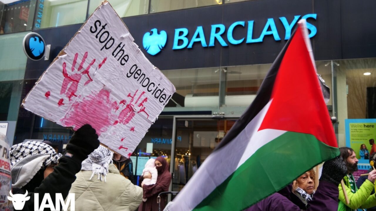 Two protesters, one wearing a keffiyeh, protest outside of a Barclays office. One holds a Palestine flag, another a sign that says "stop the genocide."