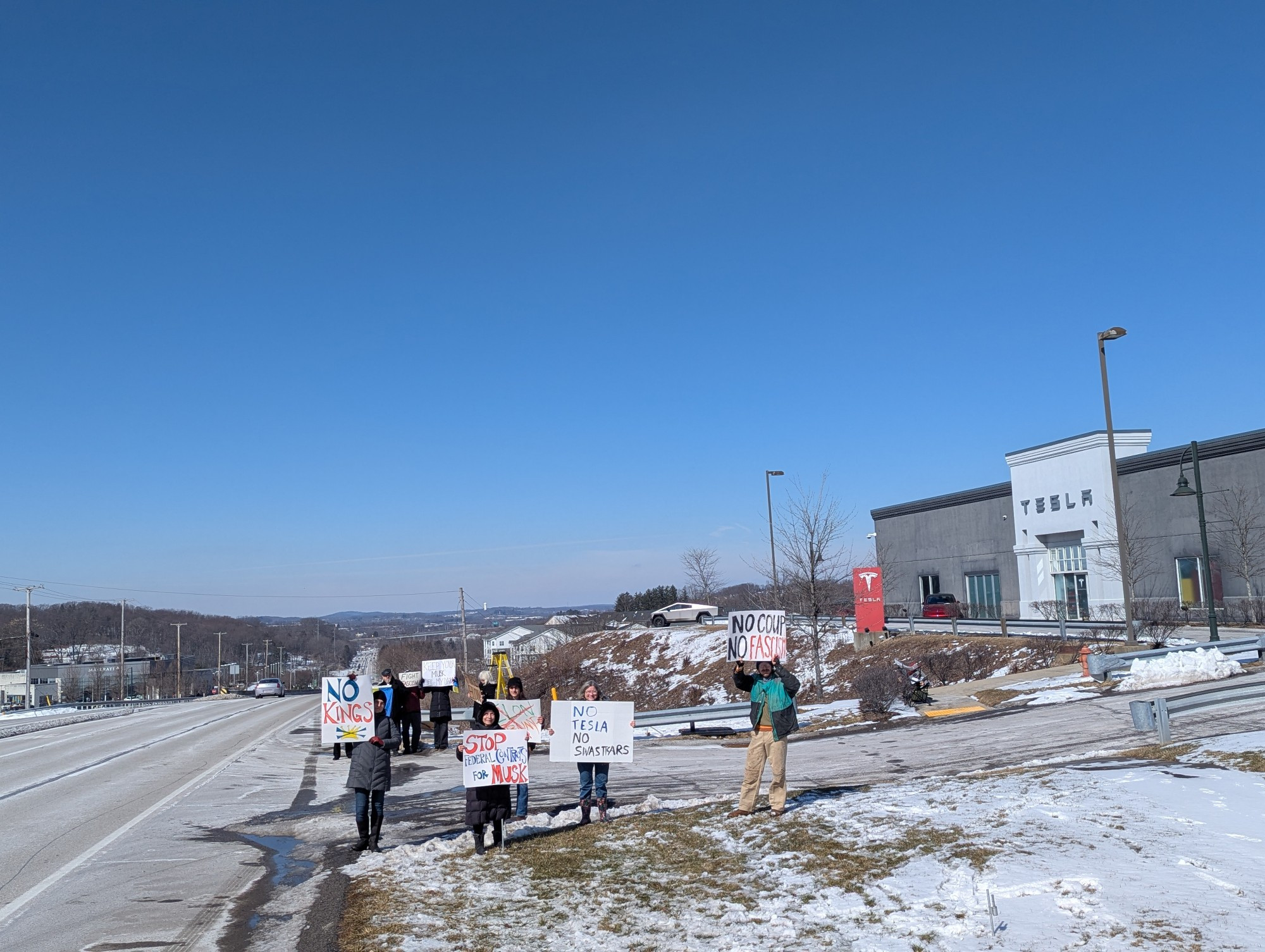 About a dozen protesters with signs, beside the road in front of a Tesla dealership.