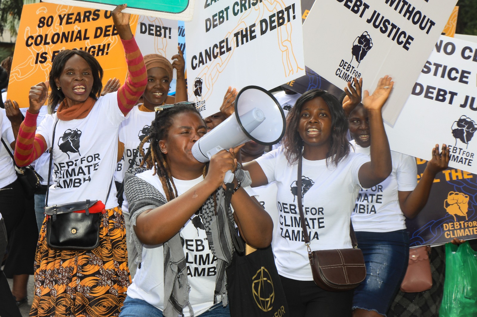 Protesters wearing 'Debt for Climate Zambia' t-shirts hold a megaphone and placards saying 'No climate justice without debt justice' and 'Cancel the debt'