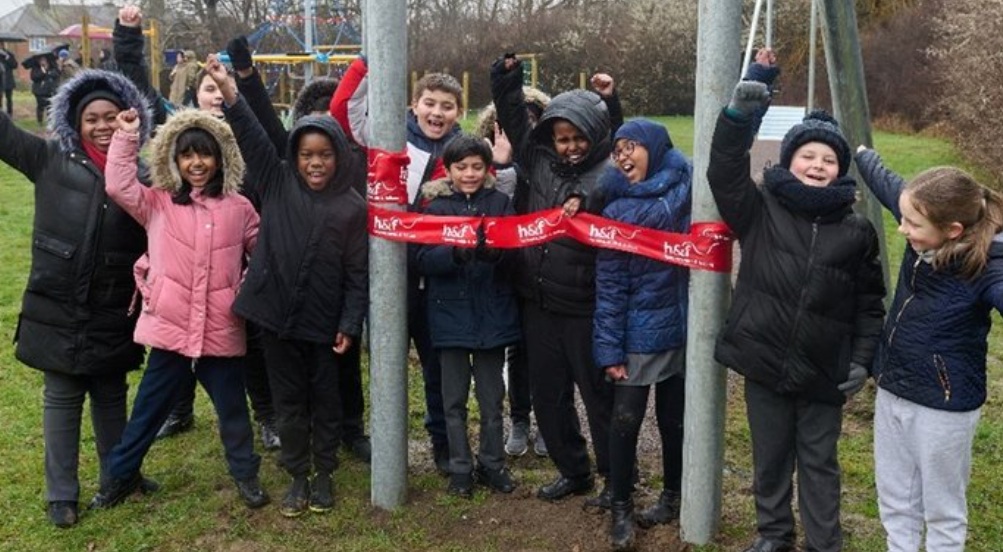 A group of children in a park standing celebrating with a red ribbon around some equipment