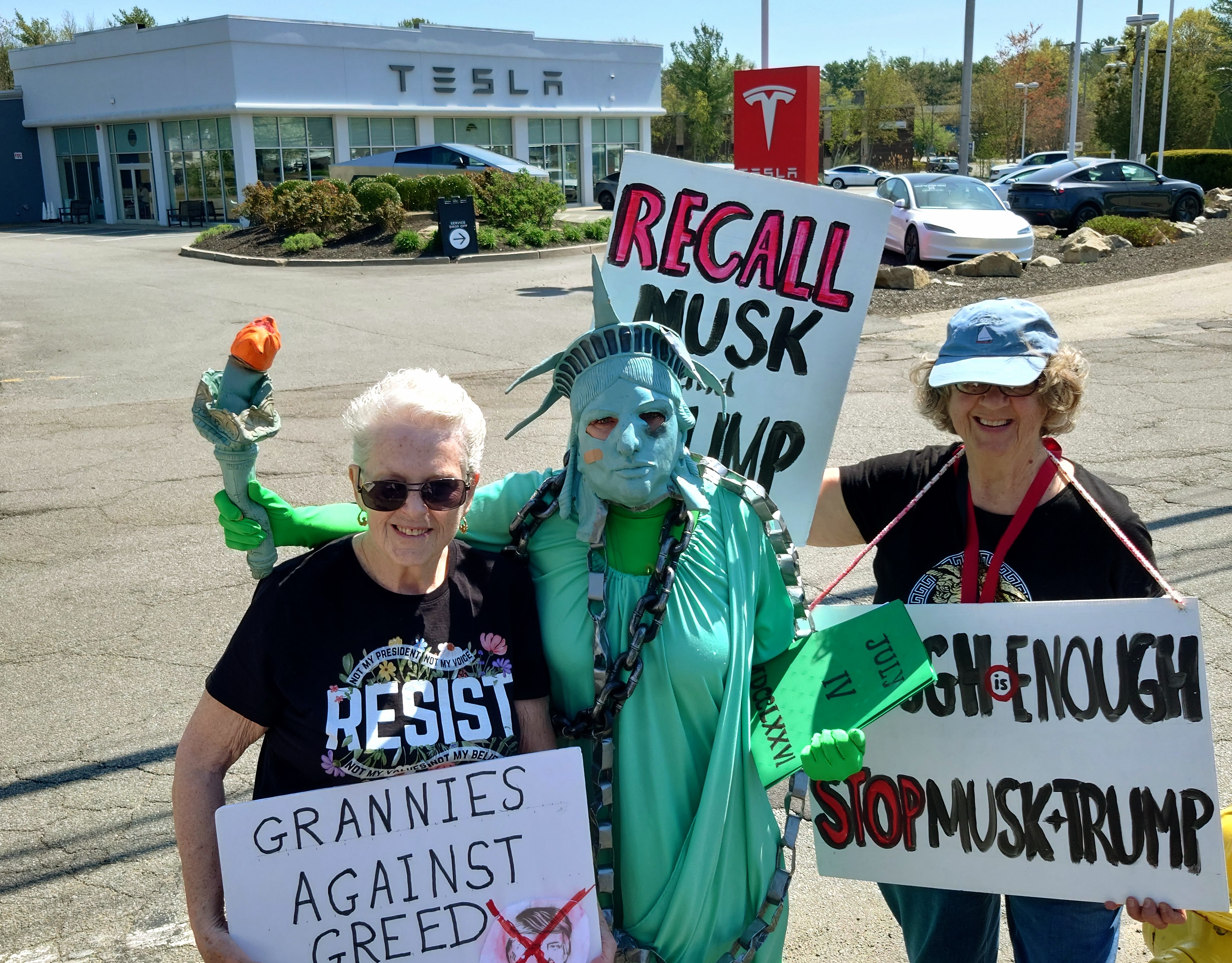 Demonstrators stand with the Statue of Liberty in chains, black eye, bandaid, crushed torch. One protestor wearing a RESIST shirt, holds a sign that reads: Grannies Against Greed. On the other side of the Lady Liberty with signs. Standing in protest in front of the Tesla Norwell MA.   