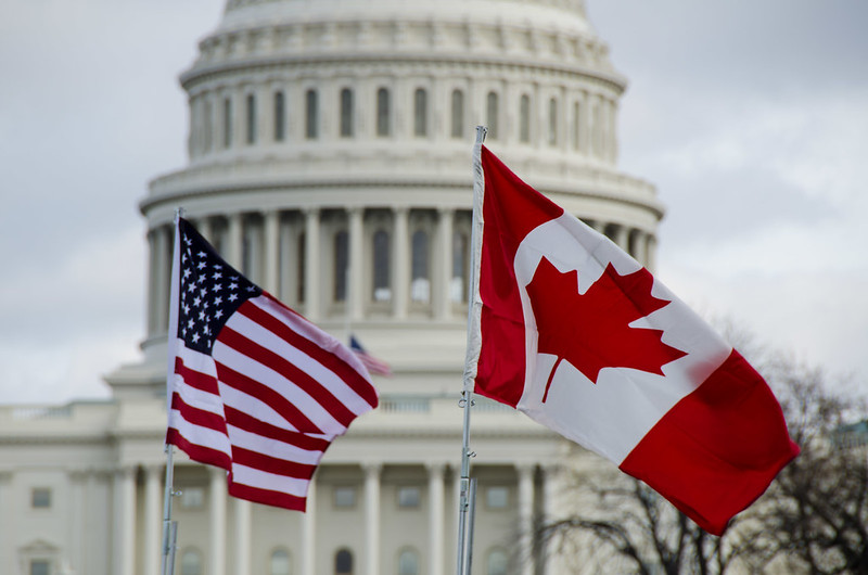 U.S. and Canadian flags fly in a blue sky in front of the U.S. Capitol