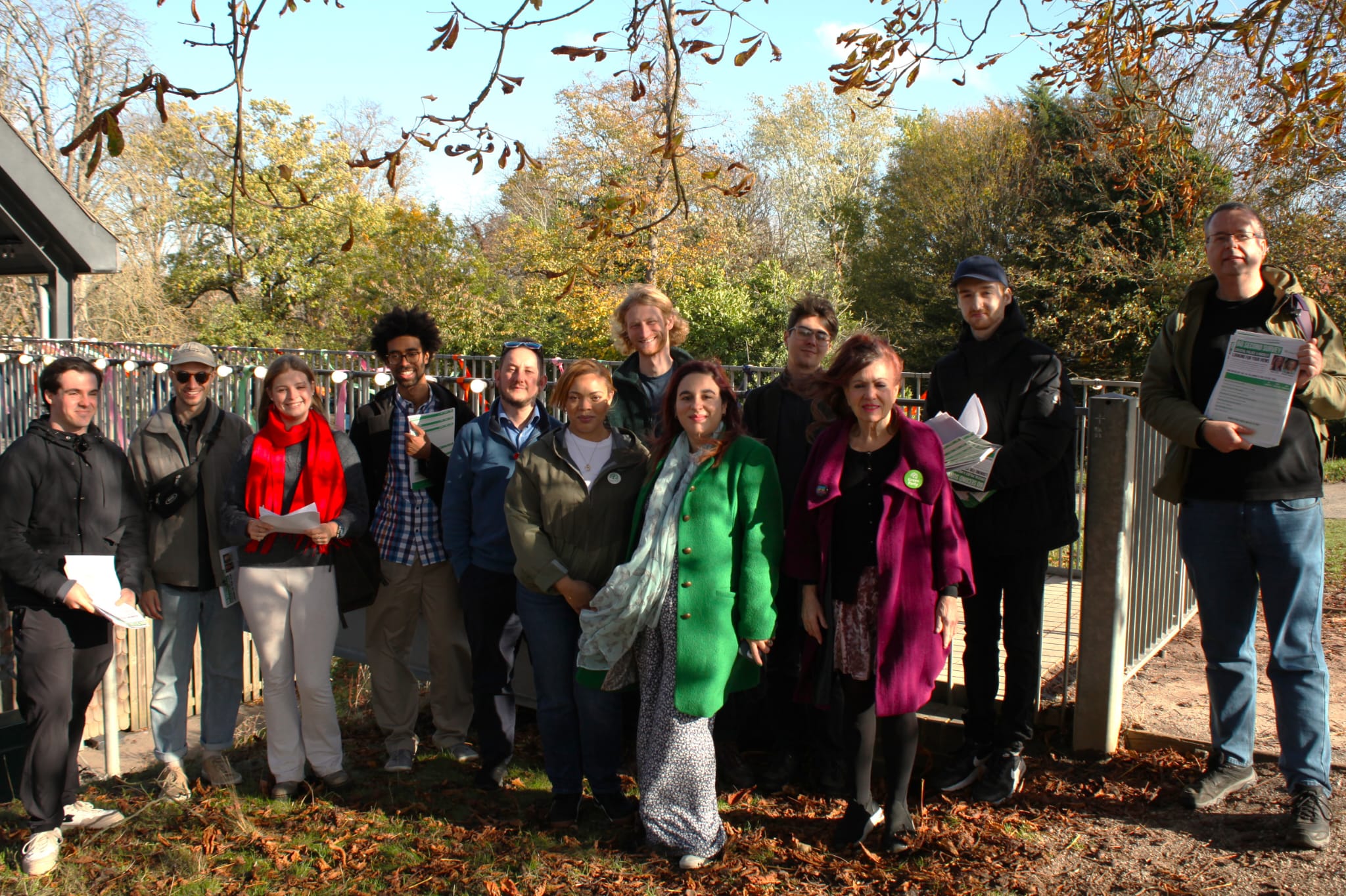 A group of thirteen people standing outdoors in autumn, smiling for a group photo. They are dressed warmly in jackets and coats, with trees and fallen leaves visible in the background. Some people are holding leaflets.