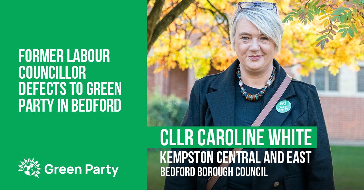 Graphic saying "Former Labour Councillor Defects to Green Party in Bedford" with Green Party logo. Then picture of Caroline. Caroline is wearing a Green Party badge on her black coat. Caroline is white and middle aged with fairly short grey hair, and her specs are up on the top of her head. Overlaid, "Cllr Caroline White, Kempston Central and East, Bedford Borough Council". In the background, grass with trees with autumnal leaves and a brick/stone building as the backdrop. 