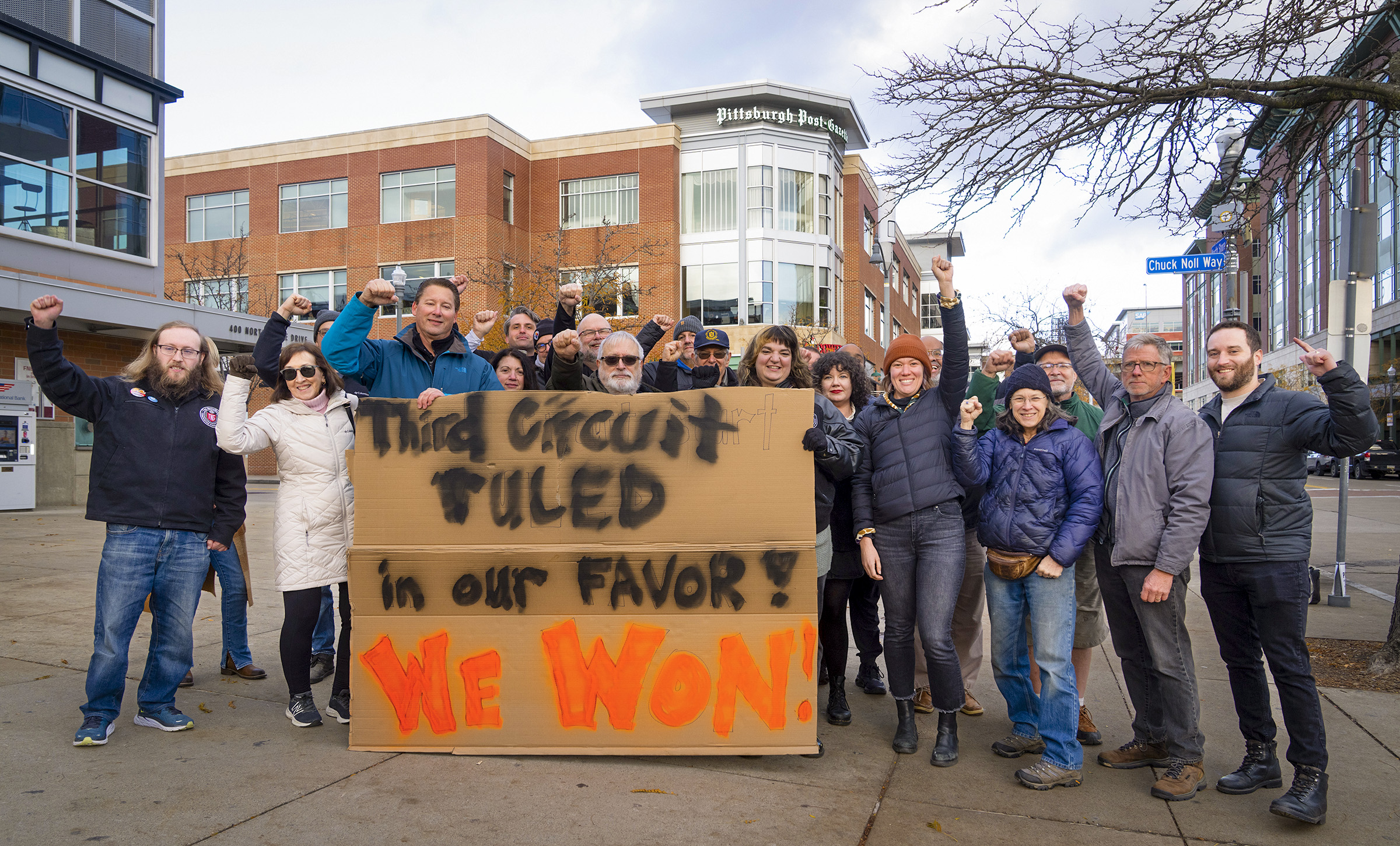 Striking workers of the Pittsburgh Post-Gazette stand behind a cardboard sign that reads "Third Circuit Court Ruled in Our Favor, We Won!" outside of Stage AE on Pittsburgh's North Shore. The struck Post-Gazette office looms in the background.