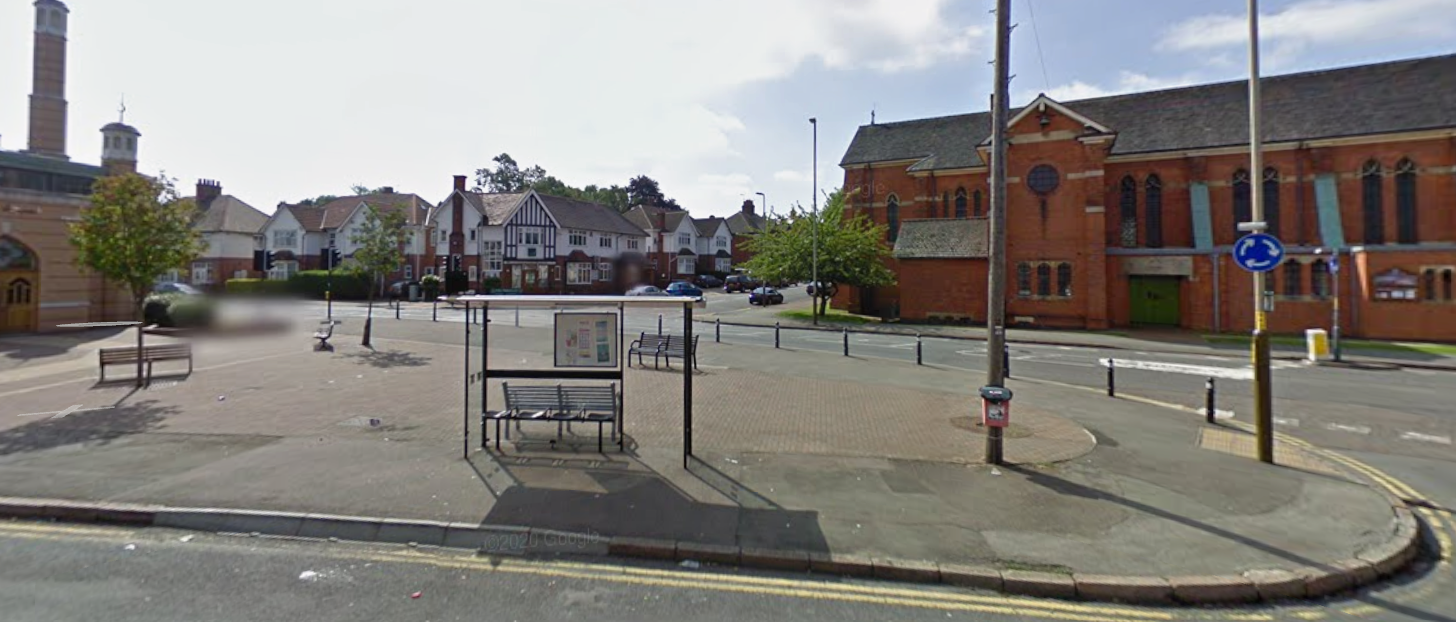 An open paved space with a bus stop and three metal benches on it. A mosque on the left, a church on the right, 1930s houses in the middle.