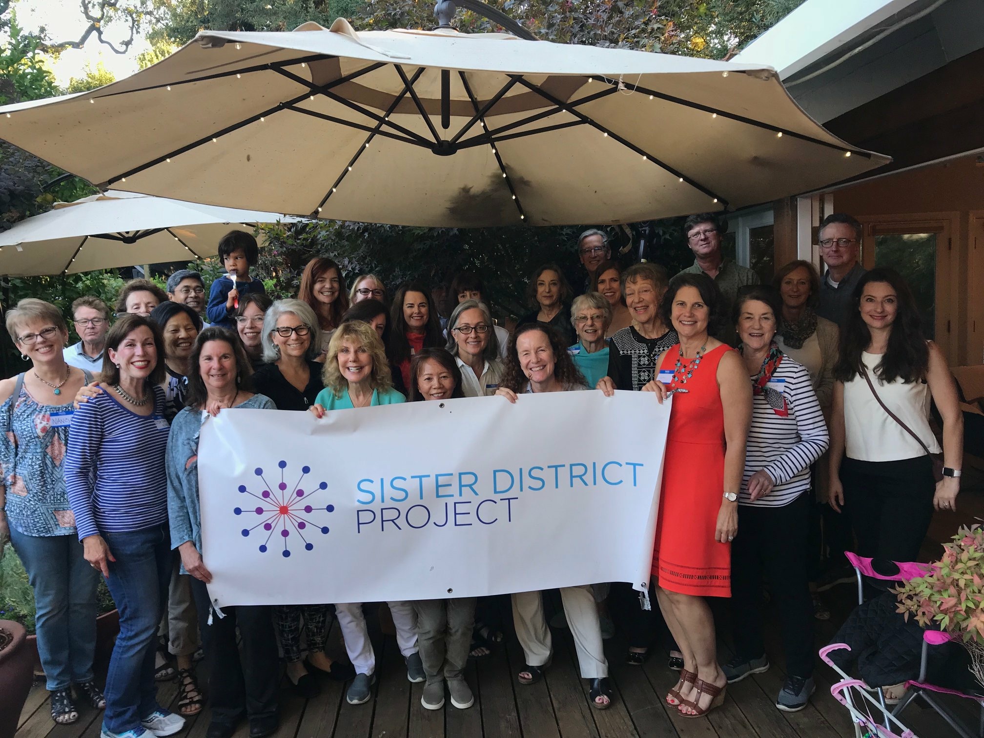 Diverse group of 30 volunteers smile at camera while those in front hold a banner reading "Sister District Project: