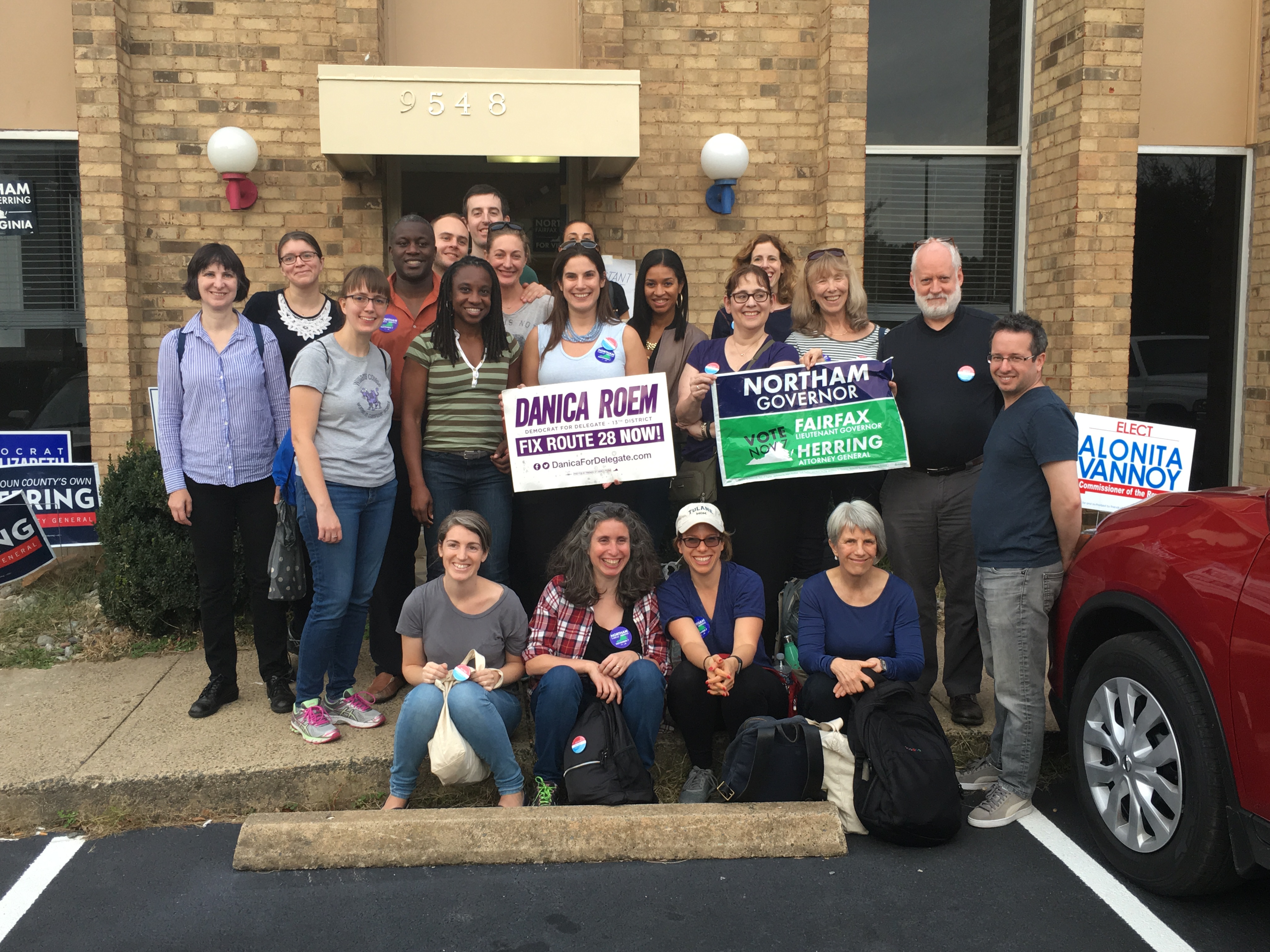 Diverse group of twenty Sister District NYC volunteers smile in front of polling location while holding signs for Virginia candidates they supported. 