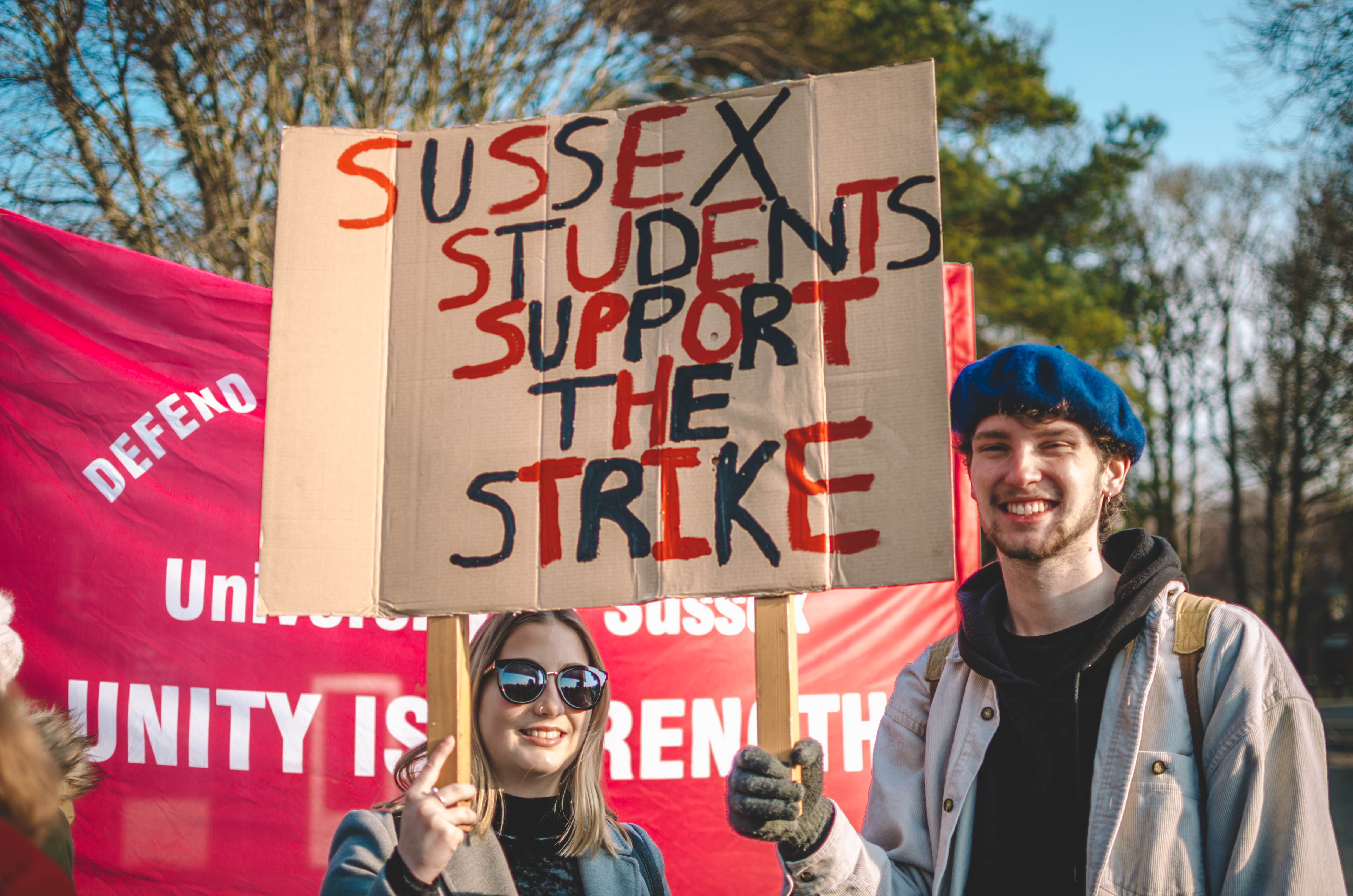 A pair of Sussex students hold a banner which reads 'Sussex Student Support the Strikes'. They are stood in the middle of a protest on Sussex Campus.