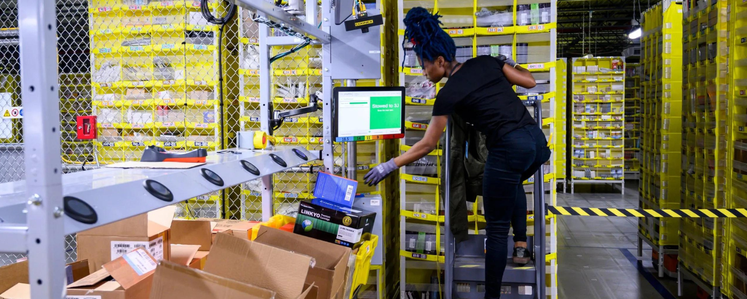 Woman in an Amazon warehouse stretching to reach a box over a convey belt.