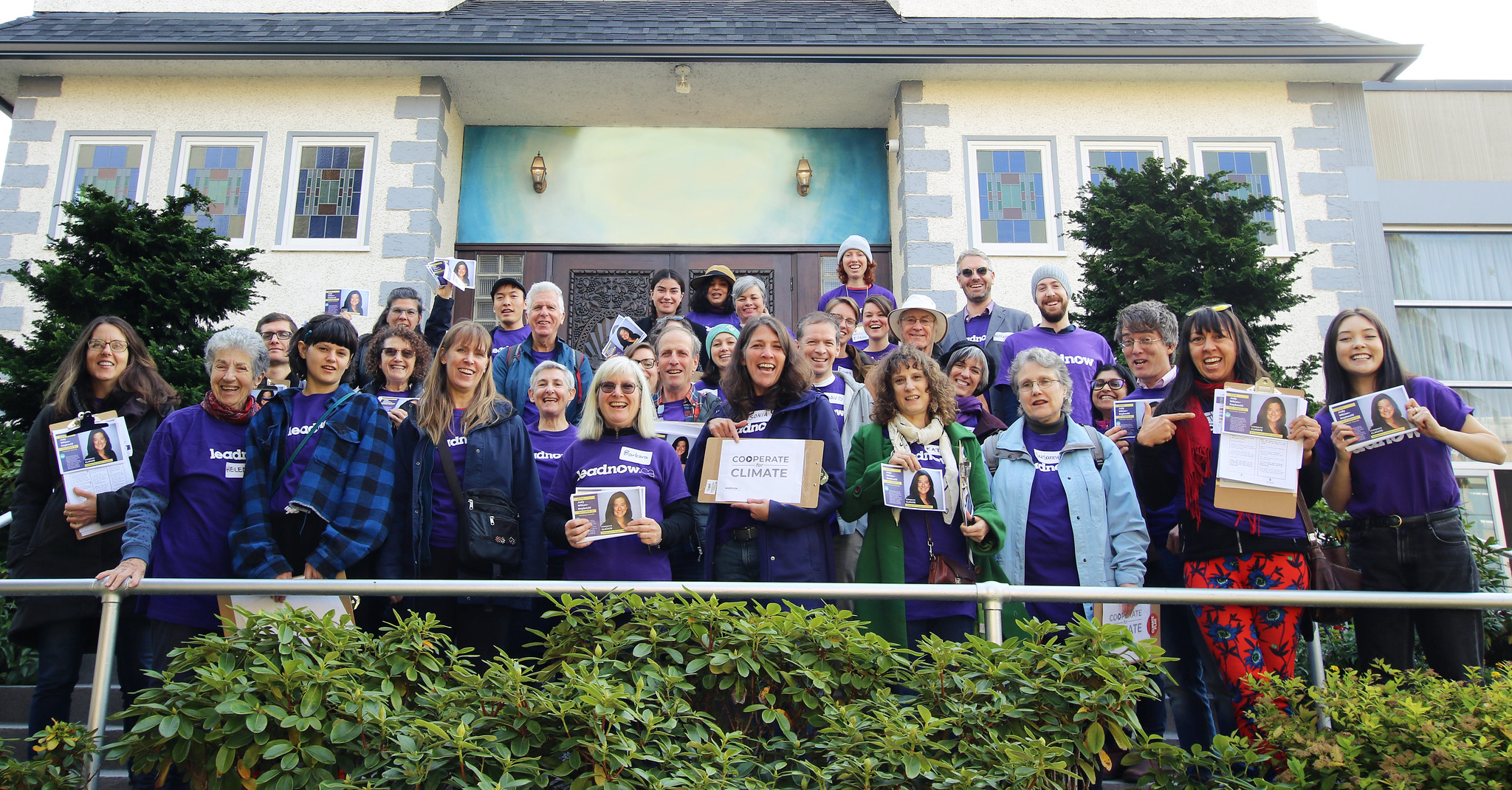Two dozen Leadnow volunteers, many wearing purple Leadnow t-shirts, hold clipboards before a doorknocking event