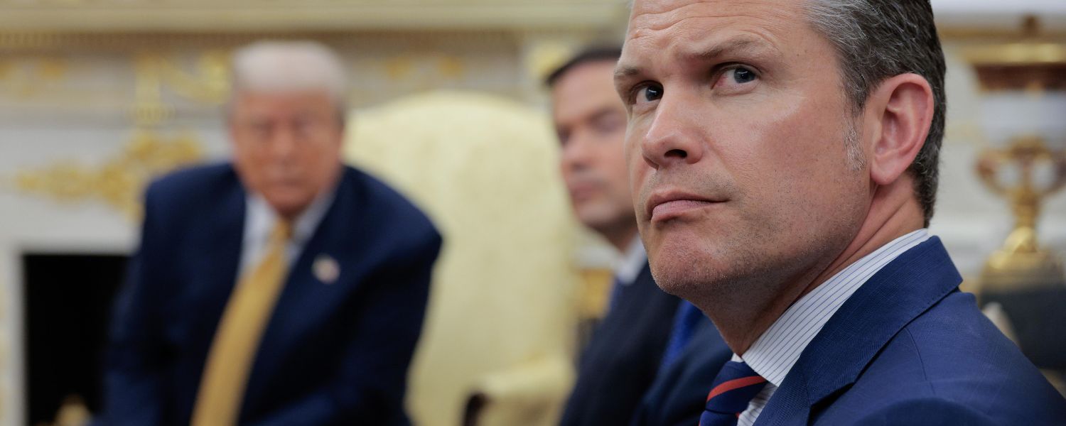 Three men in suits sit in a formal room; the man in the foreground looks to the side while two others are blurred in the background.