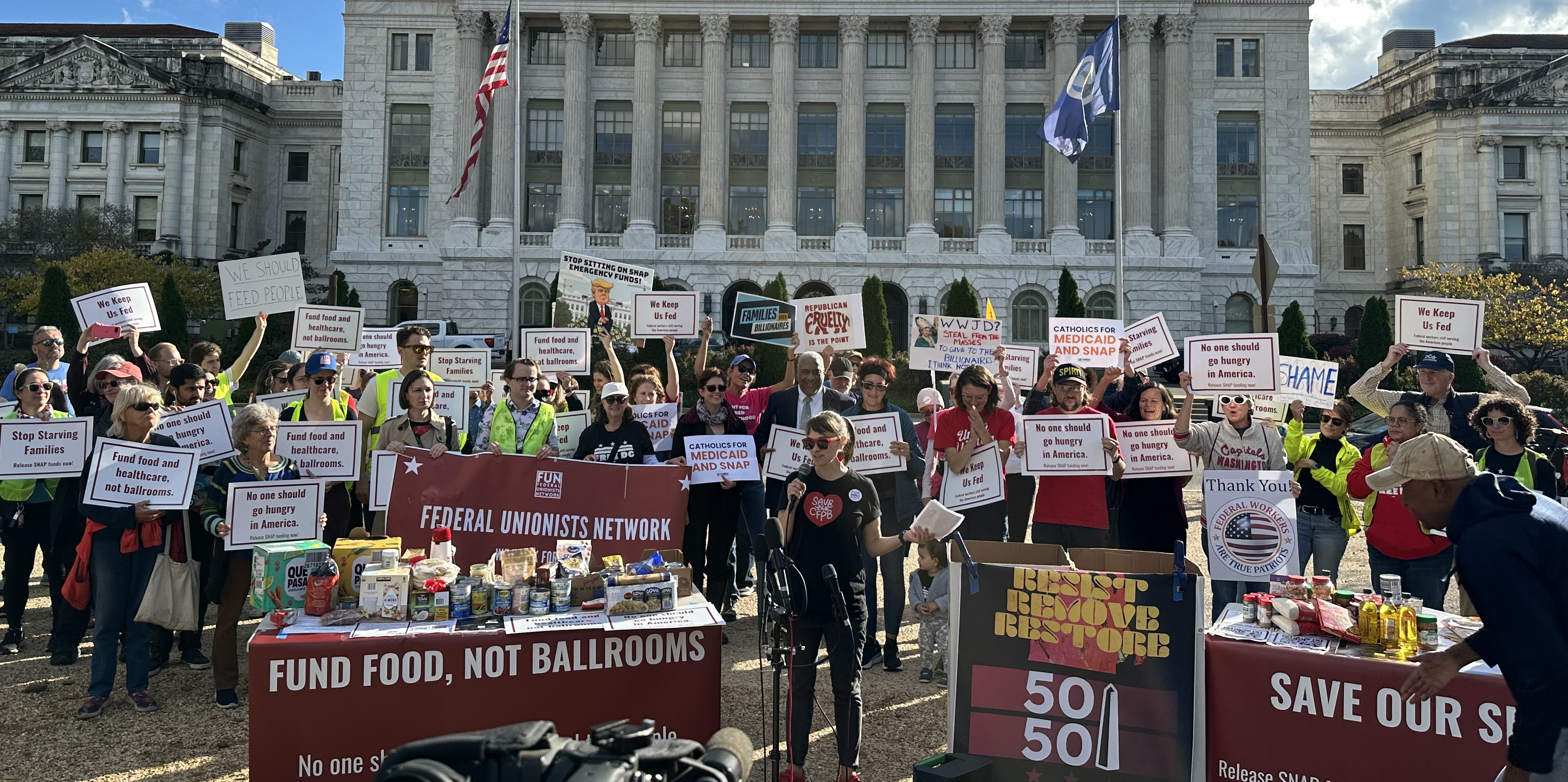 Alexis Goldstein speaks at a mic before a crowd with signs that read "fund food, not ballrooms" in front of the U.S. Department of Agriculture. Alexis is wearing a "save the CFPB" t-shirt with a redheart and red sunglasses