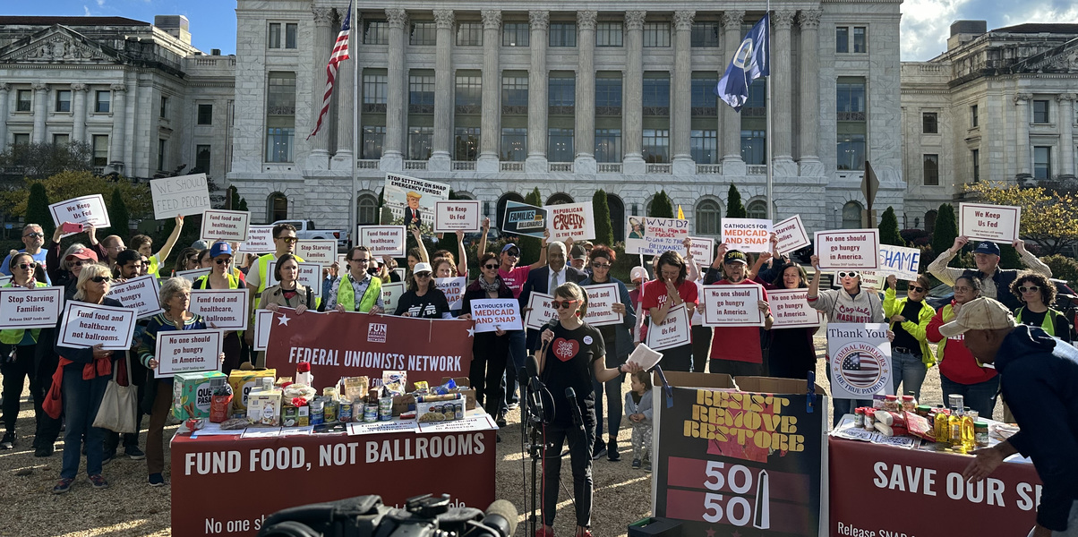 Alexis Goldstein speaks at a mic before a crowd with signs that read "fund food, not ballrooms" in front of the U.S. Department of Agriculture. Alexis is wearing a "save the CFPB" t-shirt with a redheart and red sunglasses