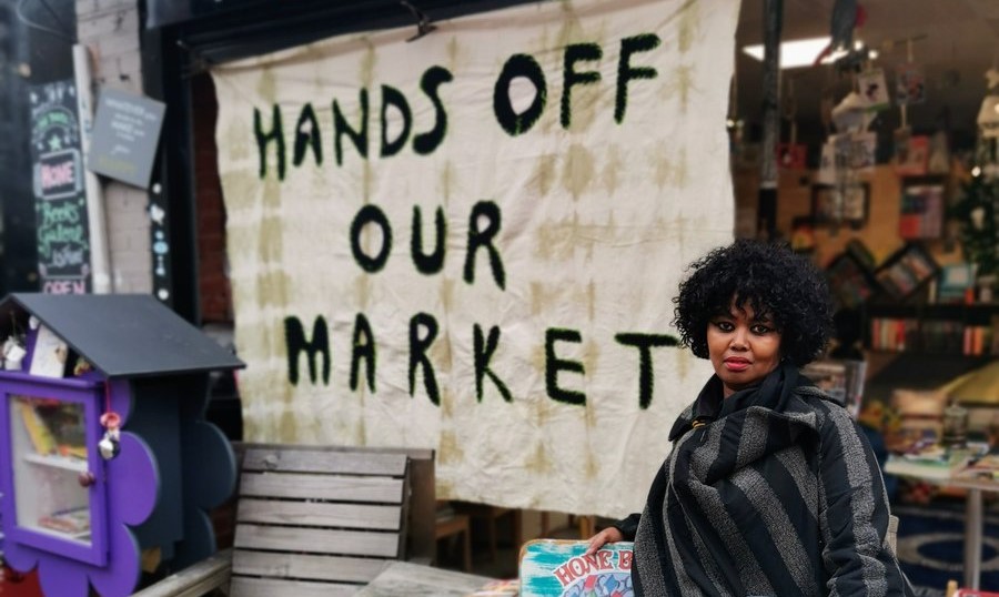 A woman stands in front of her shop, Hone Books Galore, with a banner saying 'Hands off our market'.