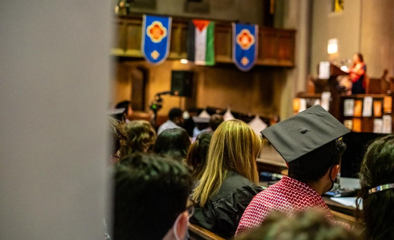 Attendees, including a graduate in a keffiyeh, sit at the People's Graduation beneath a Palestinian flag