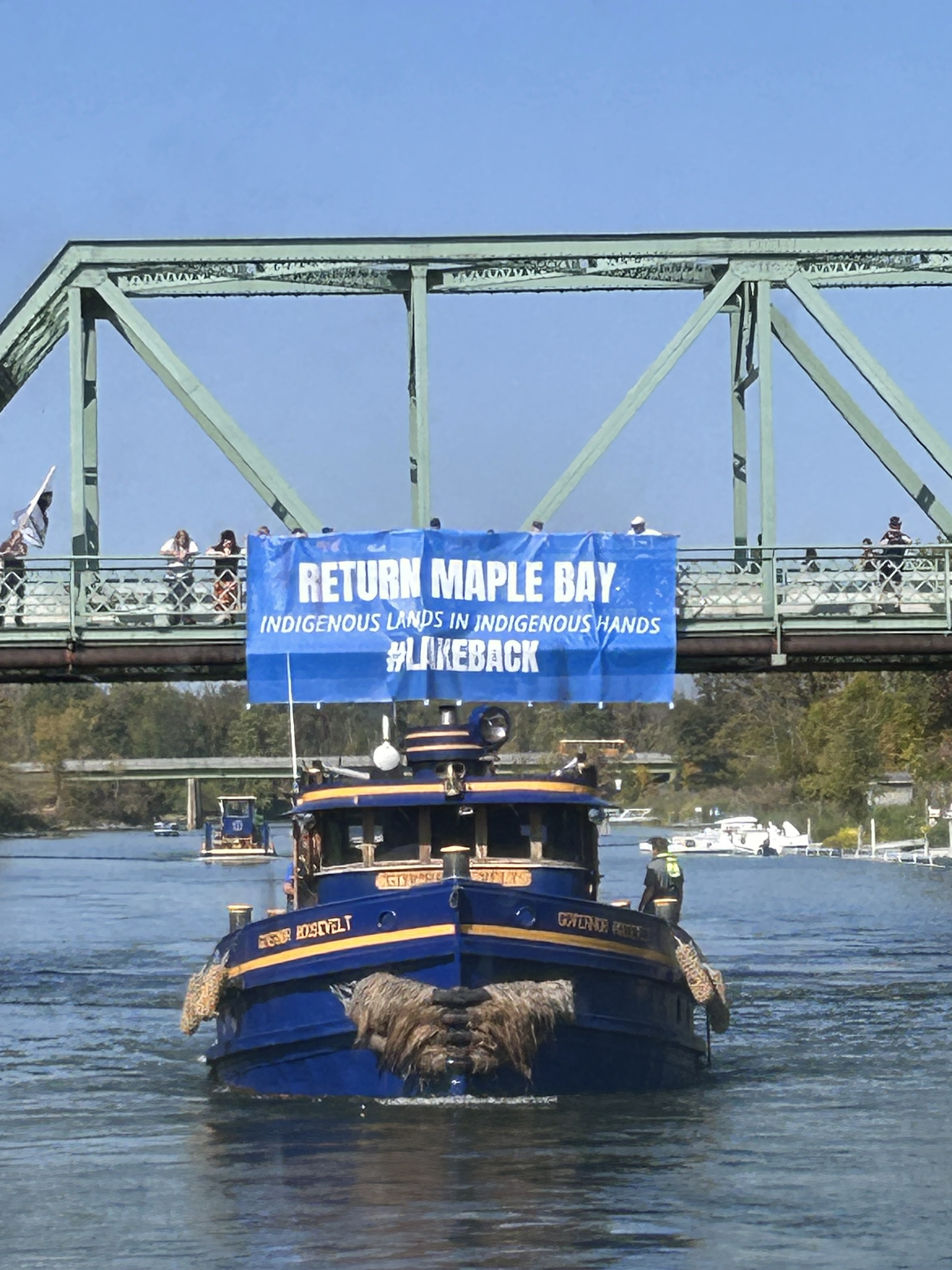 Image of a blue #lakeback banner against a green bridge as a tug boat goes underneath