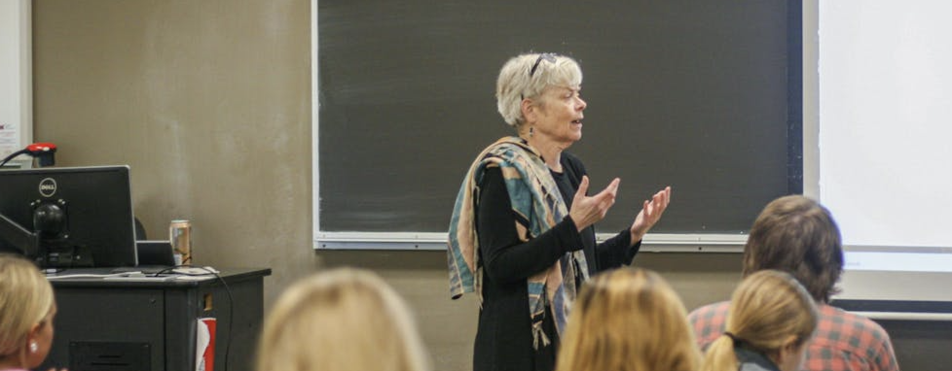 Image of a classroom with a teacher station at left, a blackboard, and attentive students listening to Professor Mary Jean Corbett teaching. Corbett is gesturing and looking off to the right of the frame at a student who is out of the picture frame.