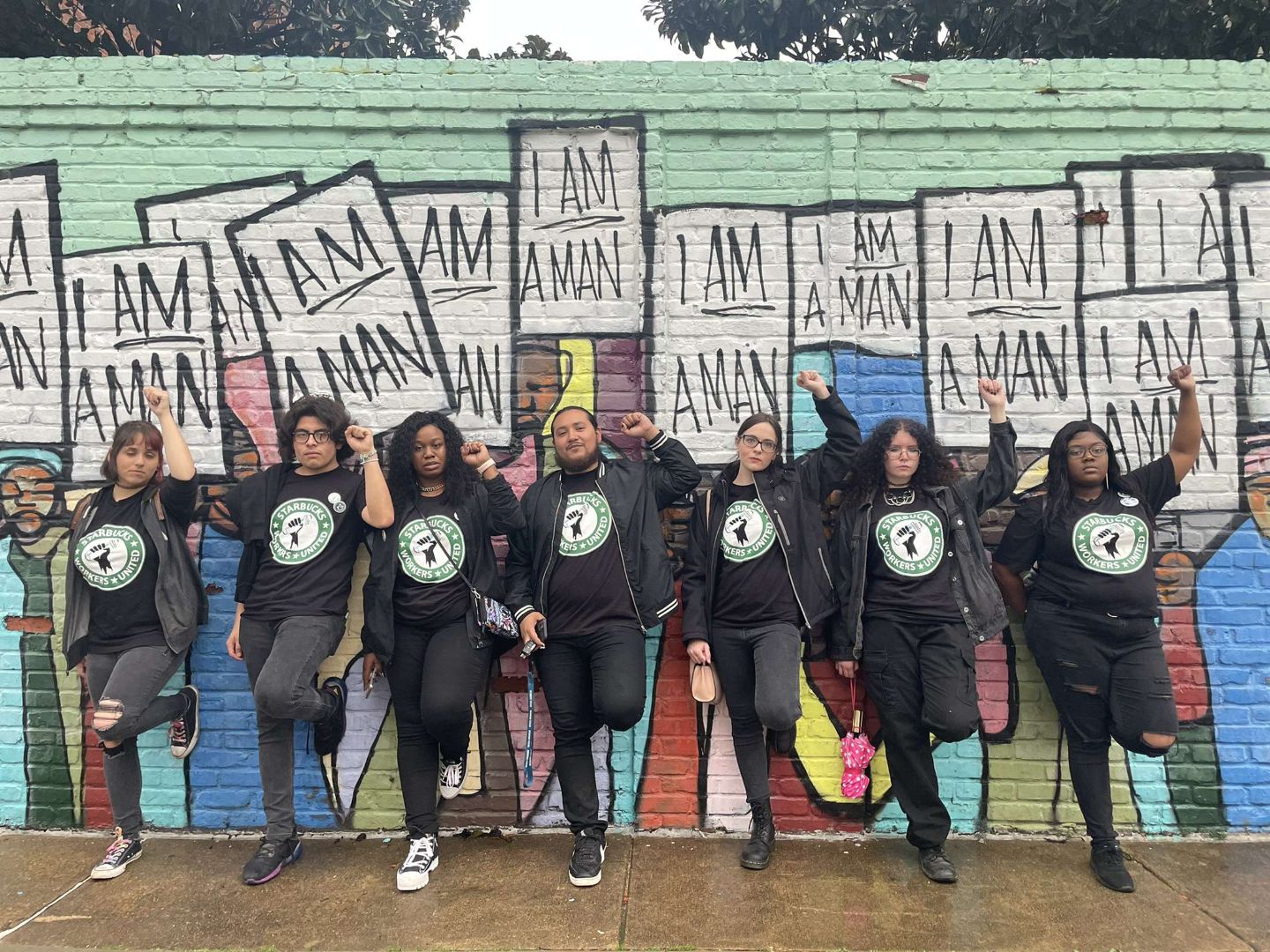 photo of seven Starbucks workers in Starbucks United tshirts, leaning against a mural wall depicting the "I am a Man" march