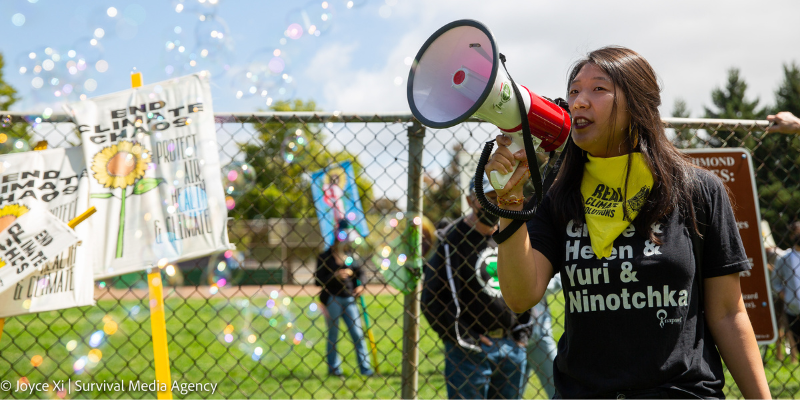 A woman holds a megaphone at a protest, surrounded by bubbles. A sign in the background reads "End Climate Chaos"