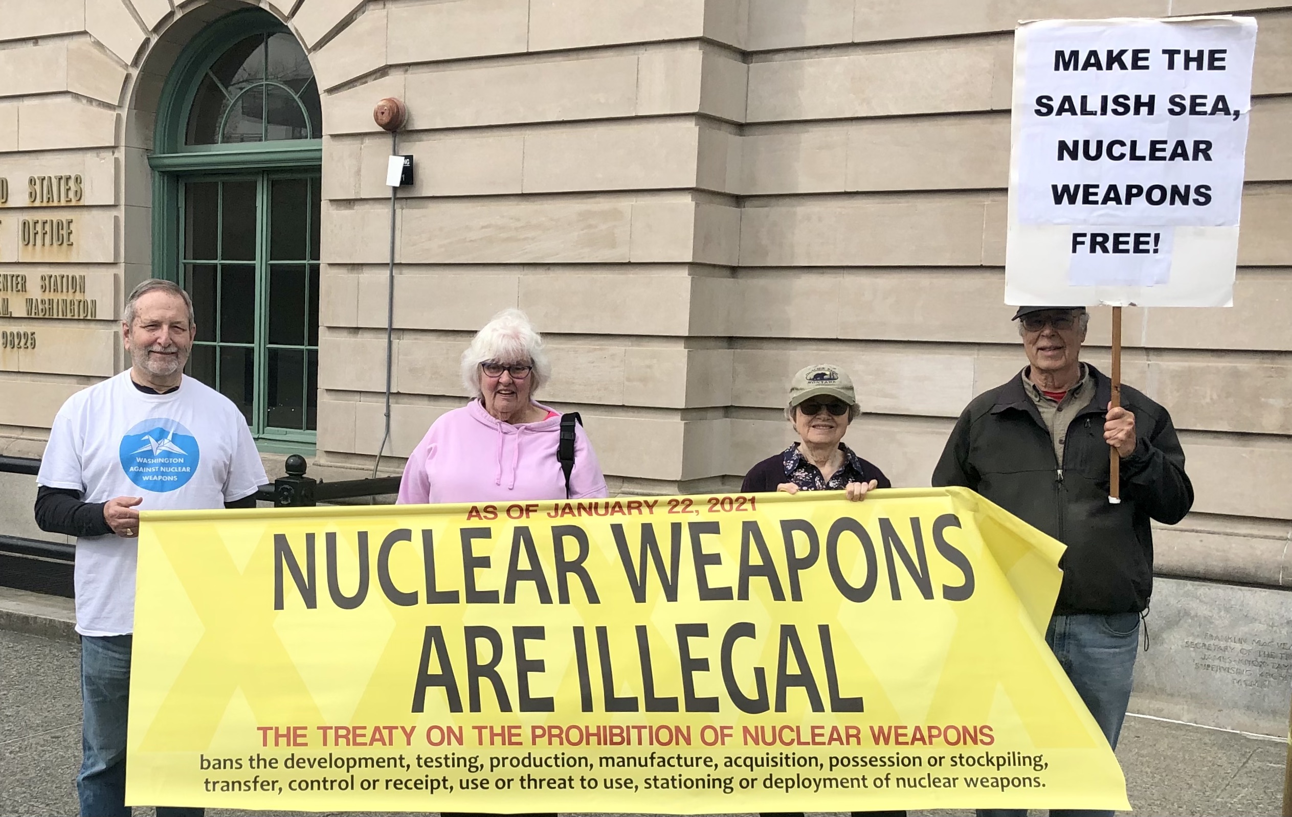 Five people hold a yellow sign in front of the Federal Building. The larger sign says:  "As of January 22 2021 Nuclear weapons are illegal - the Treaty on the Prohibition of Nuclear Weapons bans the development, test, production, manufacture, acquisition,