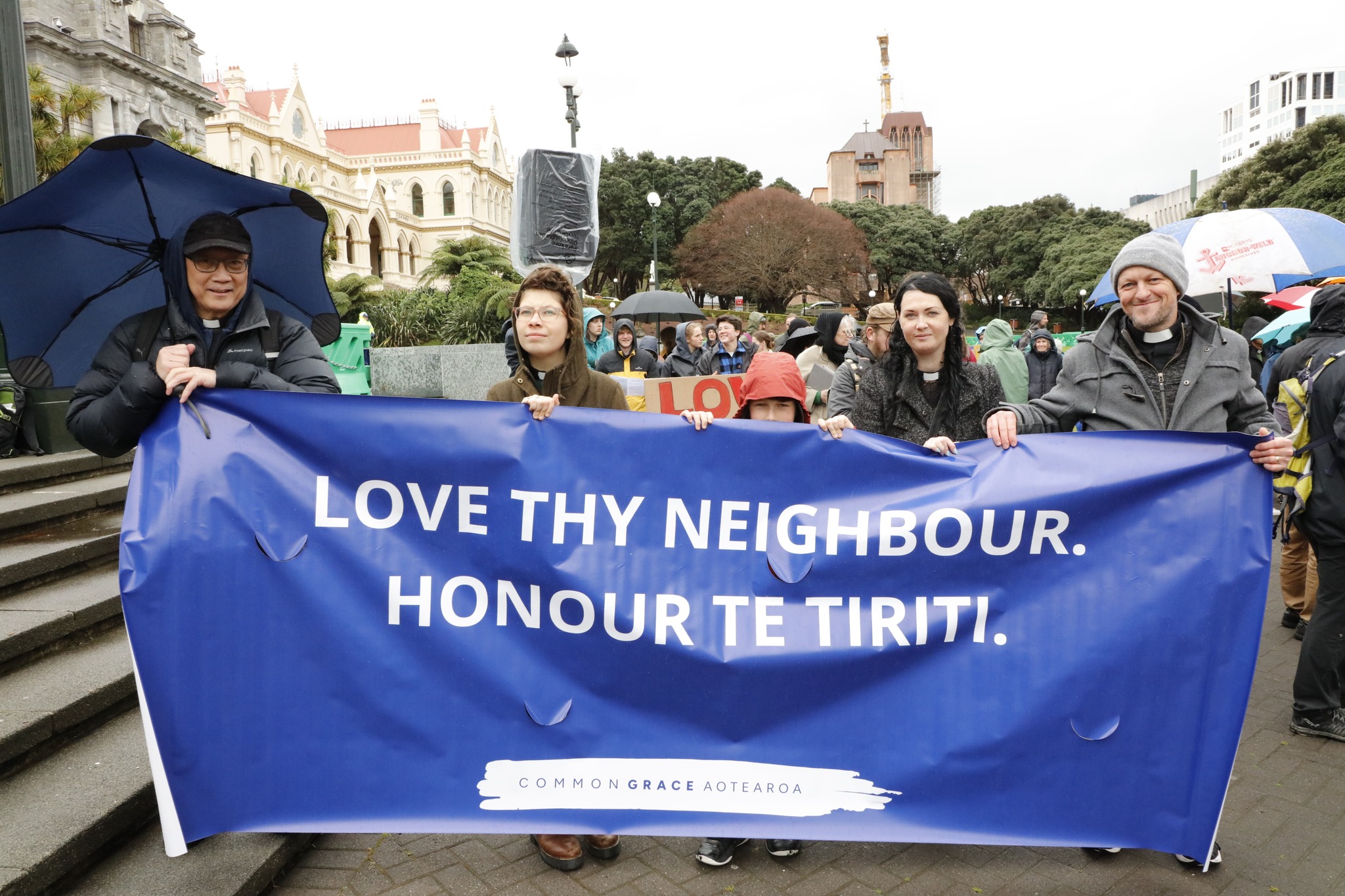 Four Anglican clergy hold a banner saying 'Love thy neighbour. Honour Te Tiriti'