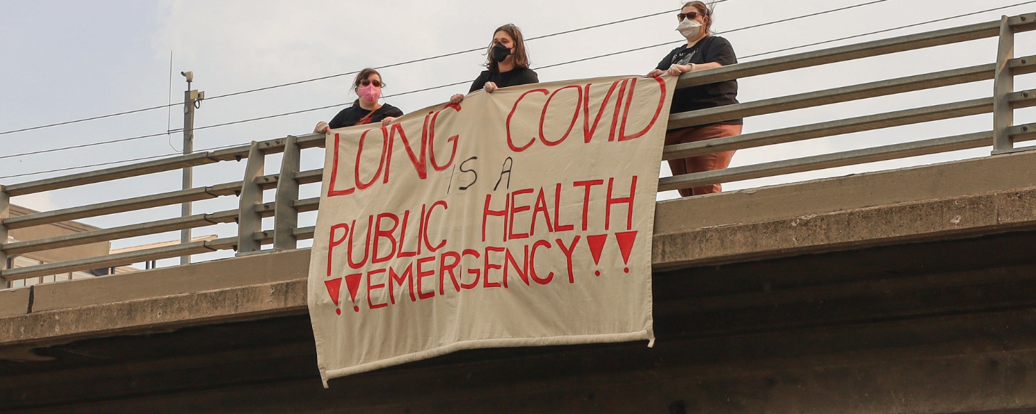 A photo of 3 masked people standing on a highway overpass with a banner that says, "Long Covid is a public health emergency!"