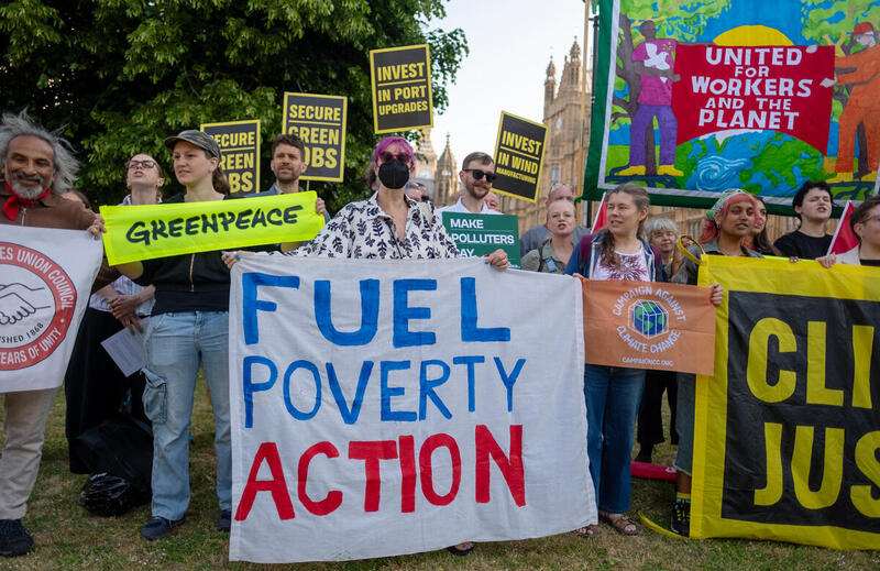 A woman holds a Fuel Poverty Action banner at a rally with Greenpeace and Campaign Against Climate Change flags either side of her.