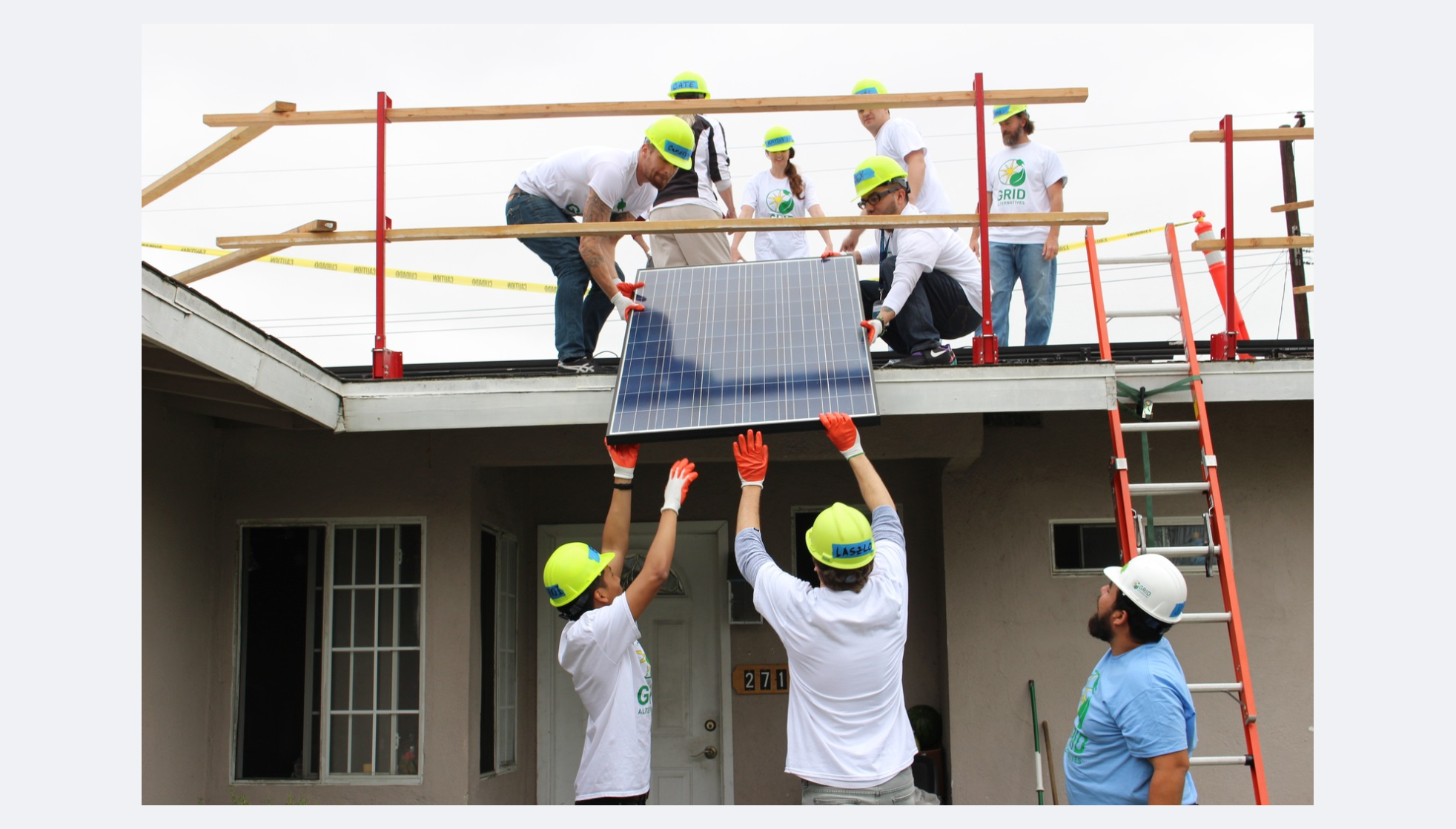 Image of people in white shirts and yellow construction hats handing a solar panel to other people standing on a roof. 