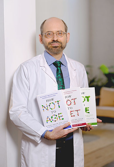 Picture of Dr. Greger holding his three books, standing in a white medical jacket in a pleasant looking lobby area