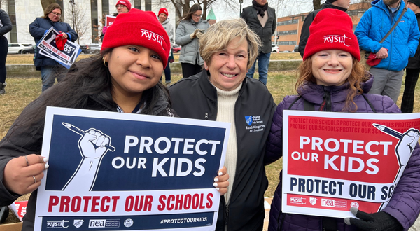 Randi Weingarten posing with protesters, with signs that say "Protect our Kids"