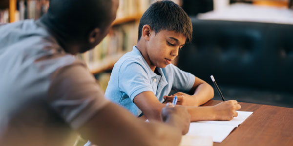 Photo of a young boy at a desk, concentrating on writing something on paper 