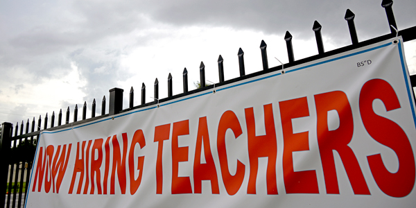 Huge banner on a fence, reading "Now Hiring Teachers"