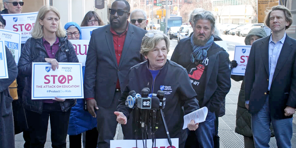 Randi Weingarten standing at a mic with protesters behind her