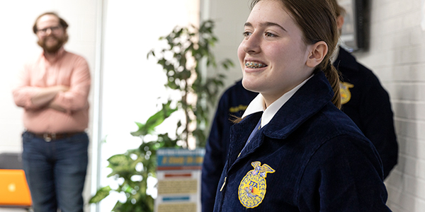 A Future Farmers of America student presents her project to a room of adults while her teacher and fellow students look on. She has long red hair and braces and is wearing a blue jacket with the FFA logo.