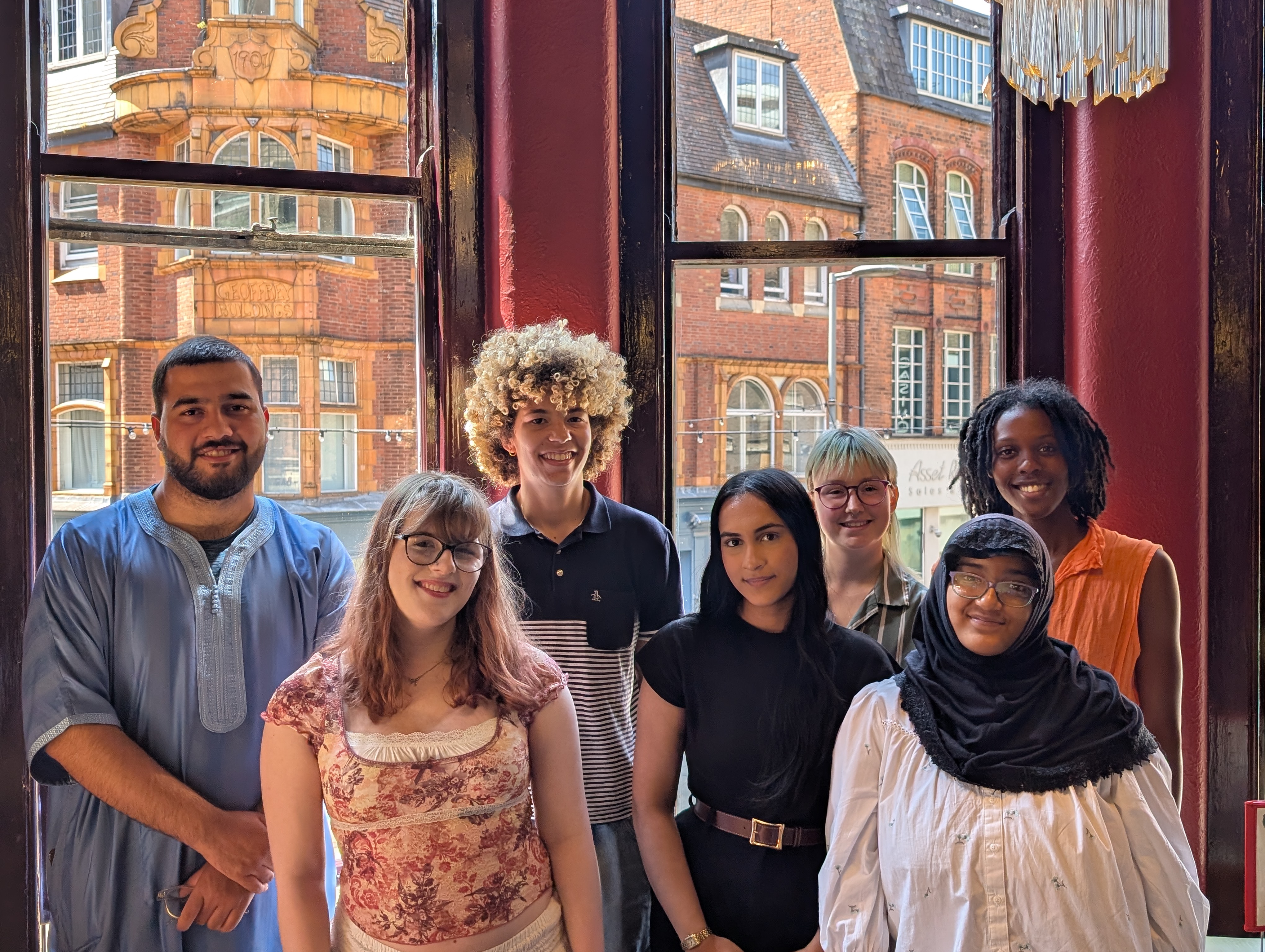 Seven members of the Youth Shadow Panel stand in front of a window, smiling