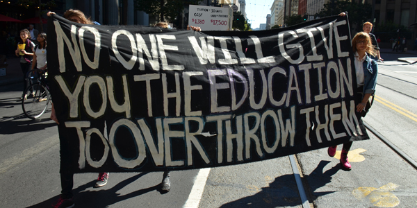 A street protest banner that says "No one will give you the education to overthrow them"
