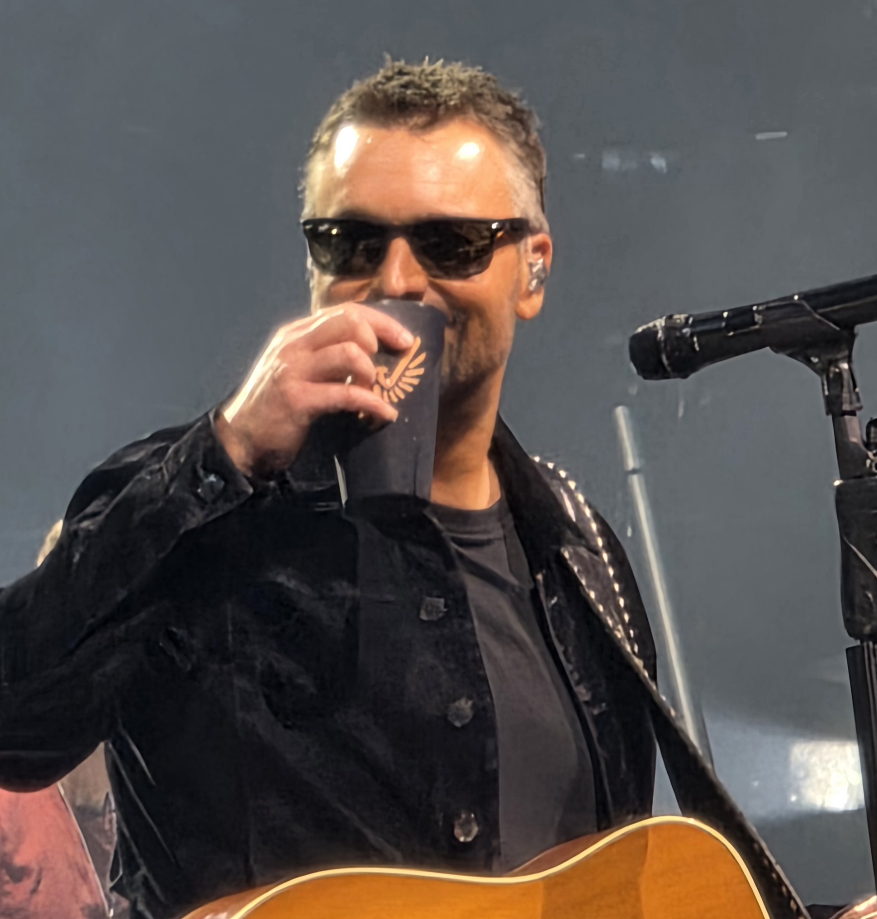 Image: Man in dark sunglasses and dark jacket, smiling, slinging a guitar and holding up his glass as if to toast 