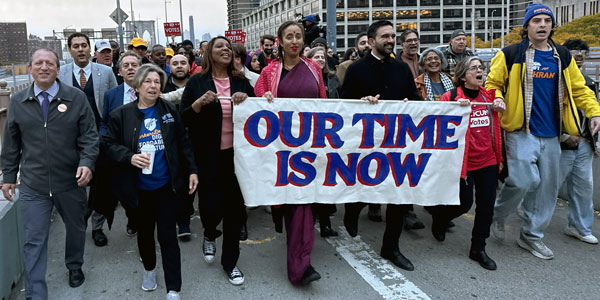 Randi Weingarten, Zohran Mamdani and a crowd of protesters with a sign, "Our Time Is Now"