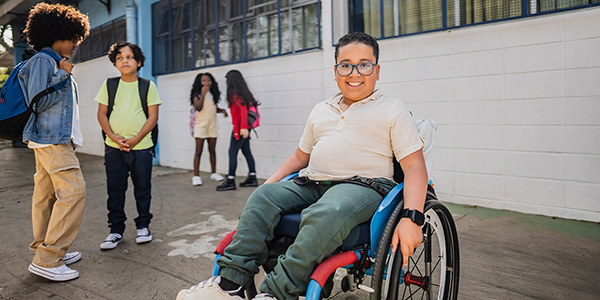 A smiling boy in a wheelchair with schoolmates standing around outdoors