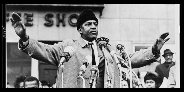 A black and white photo of Bayard Rustin with his arms out, standing in front of several microphones outdoors addressing a crowd
