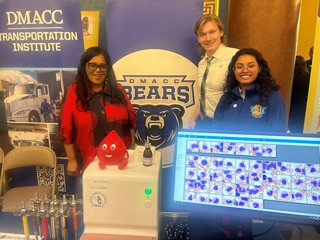 Senator Hardman with two DMACC students in the Medical Laboratory Technology program, standing behind a blood testing machine and in front of a DMACC Bears banner, during Community College Day on the Hill.