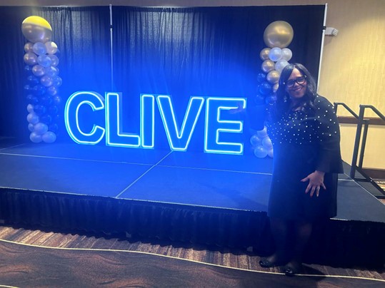 Senator Hardman stands beside a neon light display that spells out Clive on a stage at the Clive Chamber of Commerce Annual Dinner.