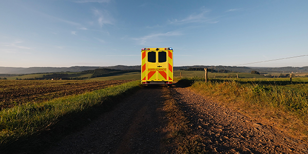 The back view of a yellow and red ambulance as it heads down a dirt road surrounded by grassy plains in the late afternoon light.