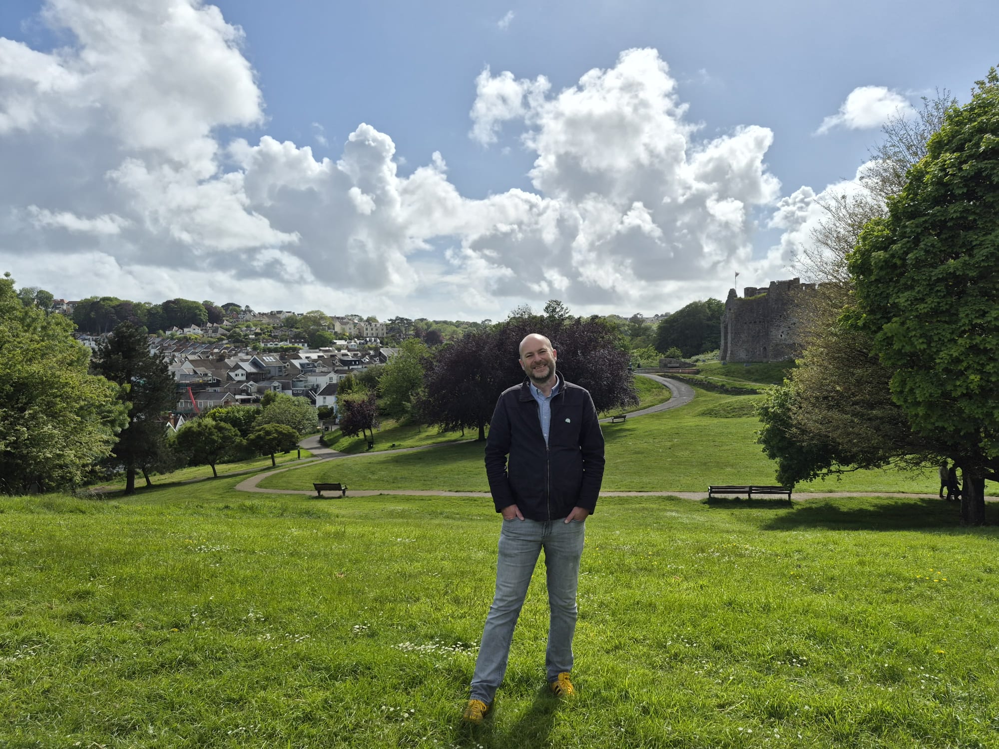 A man "Rich Williams" standing on a grassy area overlooking Mumbles and a castle in the background and a blue cloudy sky. A man "Rich Williams" standing on a grassy area overlooking Mumbles and a castle in the background and a blue cloudy sky.