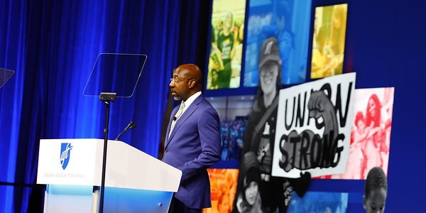 The Rev. Raphael Warnock, U.S. Senator from Georgia, stands at a podium in front of a backdrop with images of AFT members and a sign that says "Union Strong" The Rev. Raphael Warnock, U.S. Senator from Georgia, stands at a podium in front of a backdrop with images of AFT members and a sign that says "Union Strong"