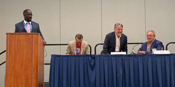 A man speaks at a podium, while three other men sit on a panel. A man speaks at a podium, while three other men sit on a panel.