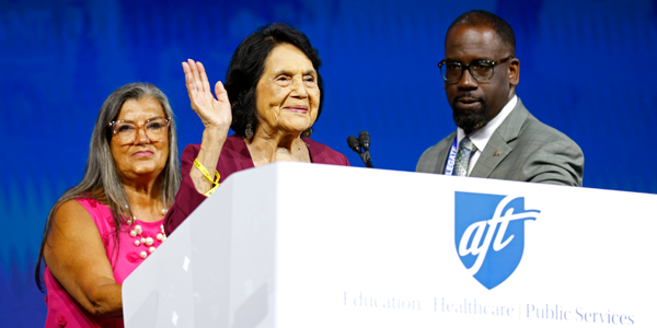 On the podium, Dolores Huerta waves to convention attendees, flanked by AFT officers Evelyn DeJesus and Fedrick Ingram. On the podium, Dolores Huerta waves to convention attendees, flanked by AFT officers Evelyn DeJesus and Fedrick Ingram.